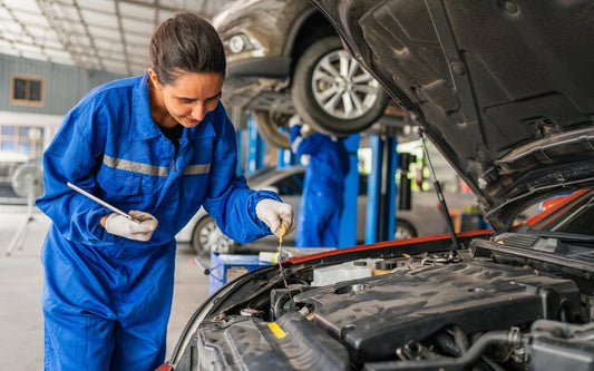 A female mechanic in a blue jumpsuit and white gloves using a tool to repair a vehicle's engine in an auto body shop.