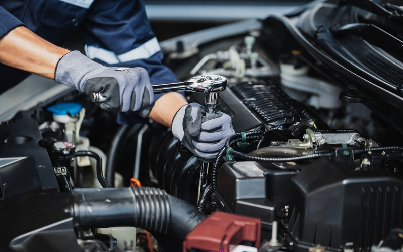 A close-up of a mechanic in a blue and silver uniform and gray gloves using silver tools to work on a vehicle engine.
