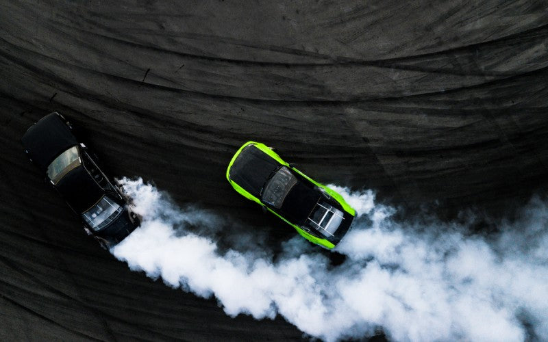 An aerial view of a black car and a black and green car racing on a black track with white smoke billowing from both cars.