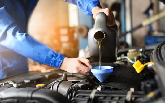 An individual in a blue jumpsuit using a blue funnel to pour amber-colored liquid into a vehicle engine.