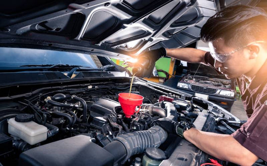 A man in safety glasses and black gloves using a red funnel to pour amber-colored liquid into a vehicle's engine.
