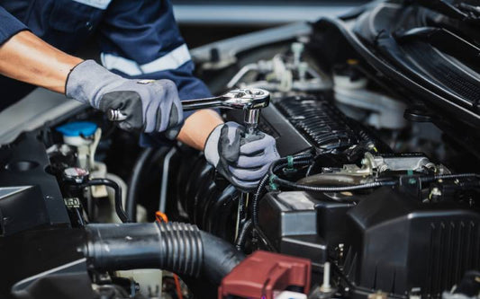 An individual in a blue and silver jacket wearing gloves while using a wrench to work on the engine of a car.