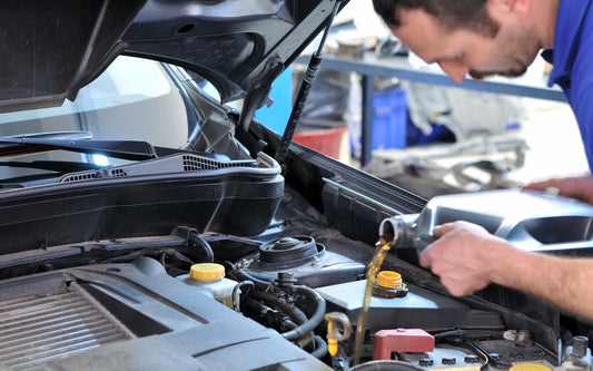 A man in a blue collared shirt holds a silver bottle and pours amber-colored oil into a vehicle's engine.