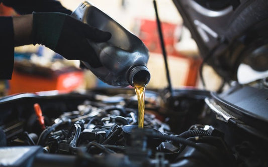 A person in black gloves uses a funnel to pour amber-colored oil from a black container into a vehicle's engine.