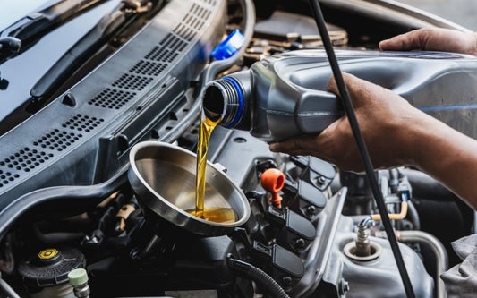 An individual uses a stainless steel funnel to pour amber-colored oil from a silver container into a vehicle engine.