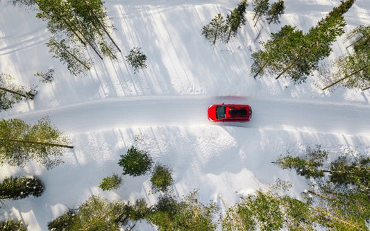 An aerial view of a red car driving on an empty, snow-covered road surrounded by tall trees on a sunny day.