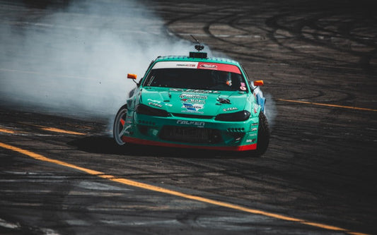 Front-facing teal drift car slides through a racetrack corner, kicking up thick tire smoke as it powers through the turn.