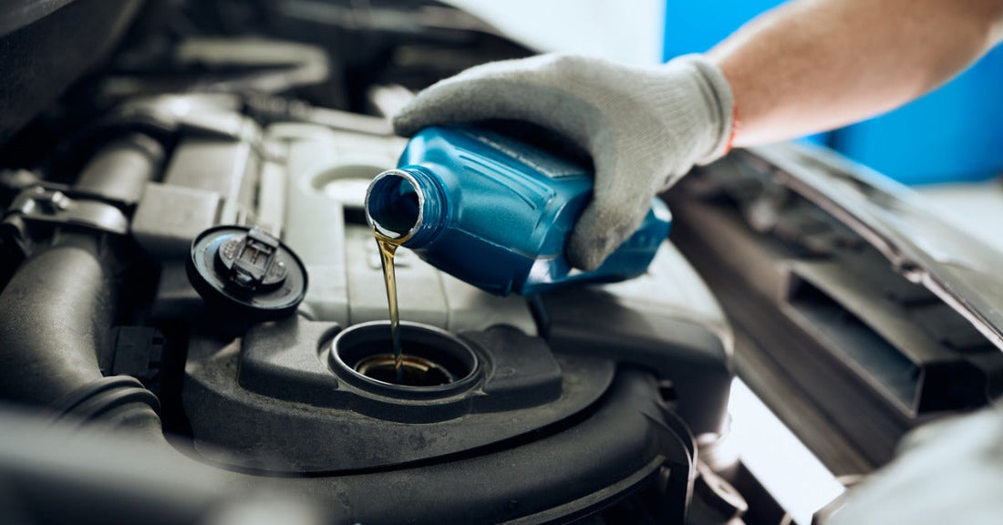 A gloved hand pours golden engine oil from a blue bottle into the oil fill opening beneath a car's raised hood.