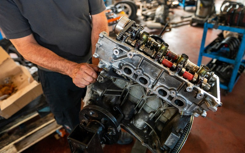 Mechanic stands beside an exposed engine cylinder head with visible valvetrain components in an automotive repair shop.