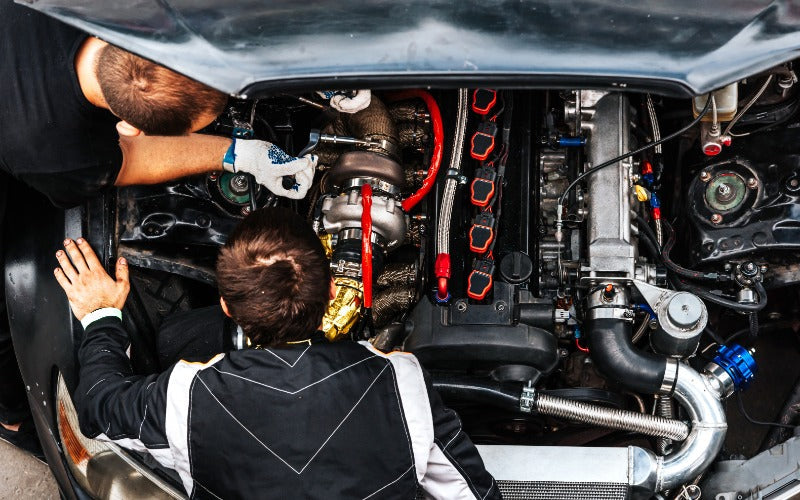 Two mechanics inspect and tune a race car engine with a large turbocharger, exposed piping, and performance components.