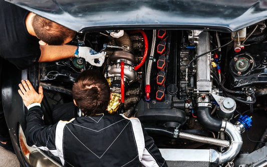 Two mechanics inspect and tune a race car engine with a large turbocharger, exposed piping, and performance components.