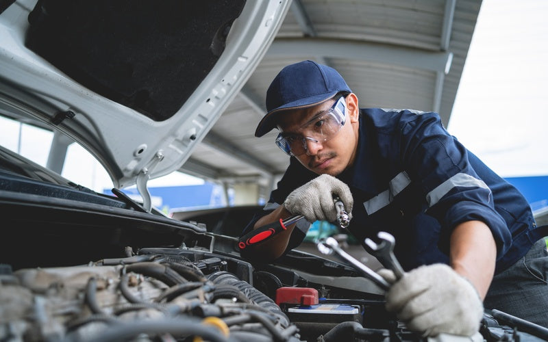 A man wearing safety glasses and gloves while holding tools and looking under the hood of a white vehicle.