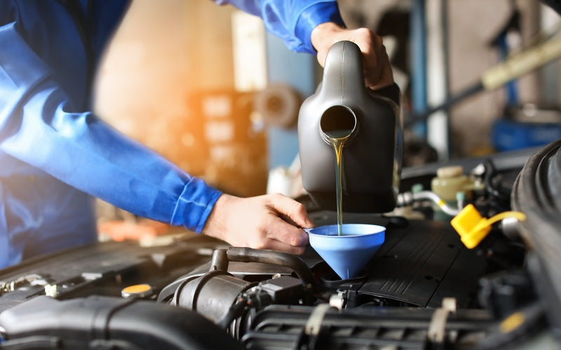 An individual in a bright blue jumpsuit using a blue funnel to pour oil into the engine of a vehicle.