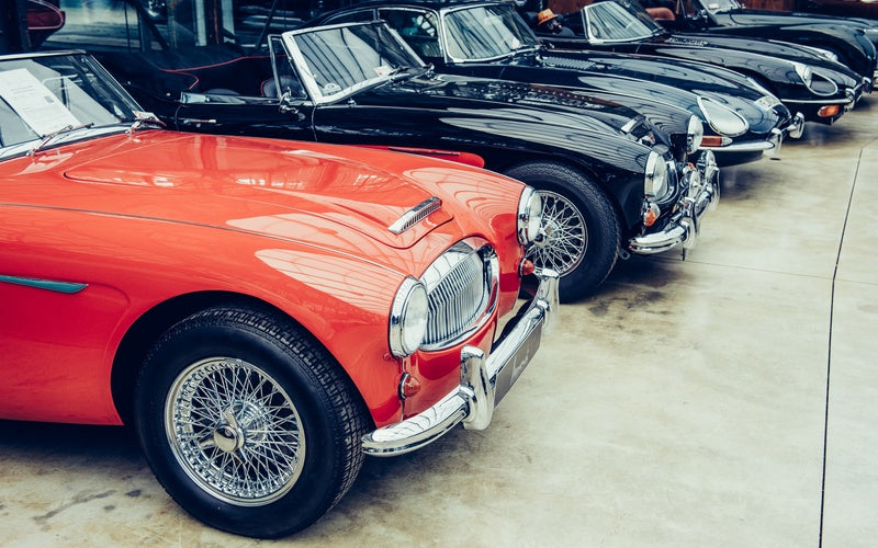 Five classic cars parked in a row on a concrete surface. The first car is red and the other four are black.