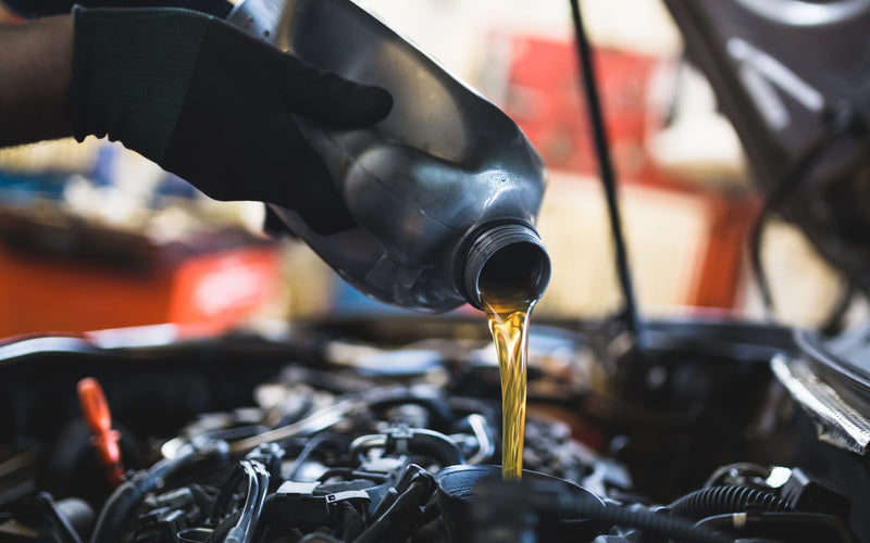 An individual wearing black gloves while pouring amber-colored liquid into a black funnel placed in a car engine.