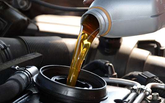 An individual pouring amber-colored oil from a silver container into a circular component of a car engine.