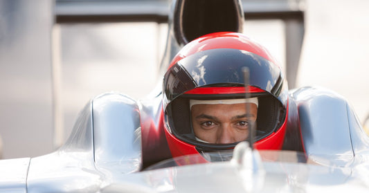 A race car driver in a white balaclava and red and black helmet sitting behind the wheel of a silver vehicle.