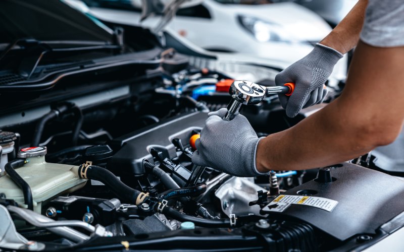 An individual in a gray T-shirt and gloves using a wrench to work on a car engine. There are other cars in the background.