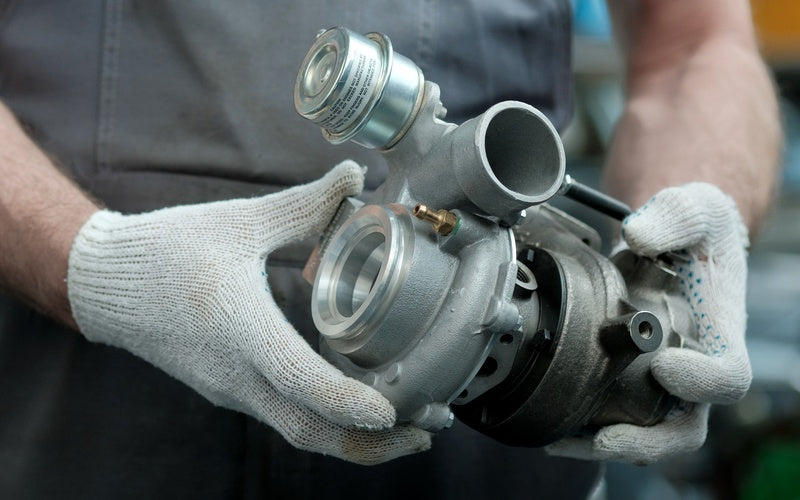 A close-up of an individual in gray clothing and white gloves holding a large silver engine in both hands.