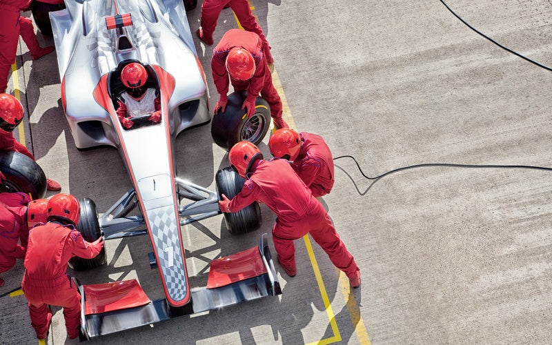 An aerial view of a crew team in red uniforms and helmets changing the tires on a red and silver race car.