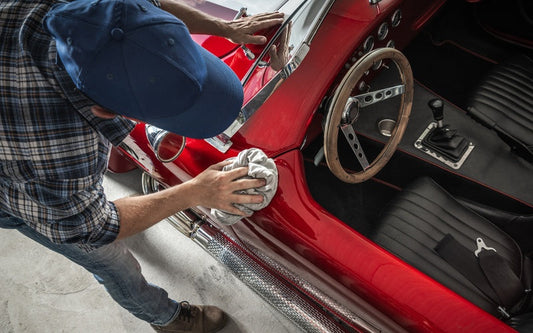 An overhead view of a man in a plaid shirt and blue hat using a cloth to polish the exterior of a red classic car.