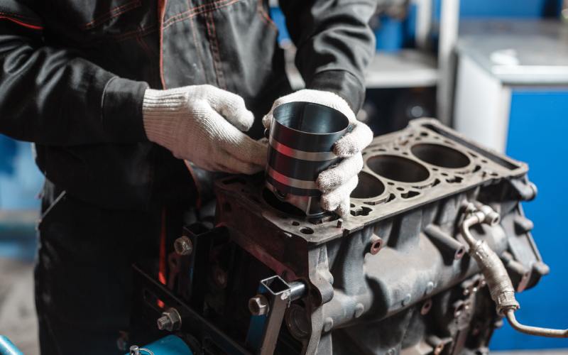 A person in a black jumpsuit and white gloves installing a black and silver piston in a disassembled engine.