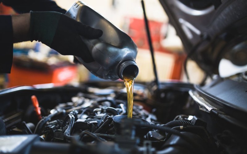 A person in black gloves uses a funnel to pour amber-colored oil from a black container into a vehicle's engine.