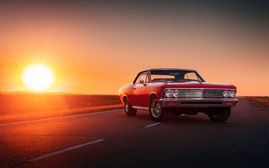 A red muscle car with chrome accents is parked on an asphalt road, next to a field, as the sun sets.