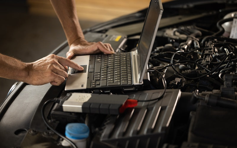 A person uses a black laptop connected to a vehicle engine to run a diagnostic test to identify potential issues.