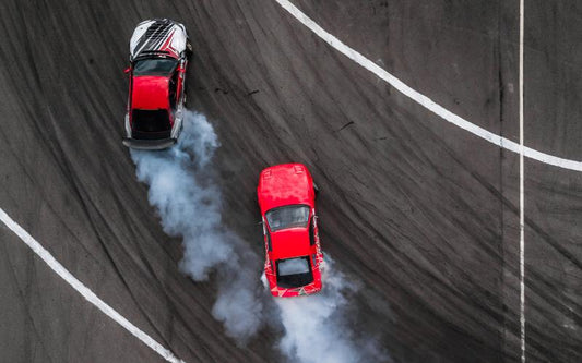 An aerial view of two race cars drifting on an asphalt track, with smoke billowing from their rear tires.