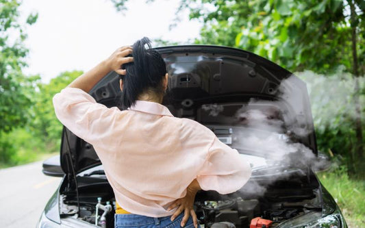 A woman in a pink shirt stands in front of her car with her hand on her head as smoke billows from the engine.