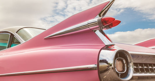 The back of a pink classic car with chrome details on its bumper and sides, parked outside on a cloudy day.