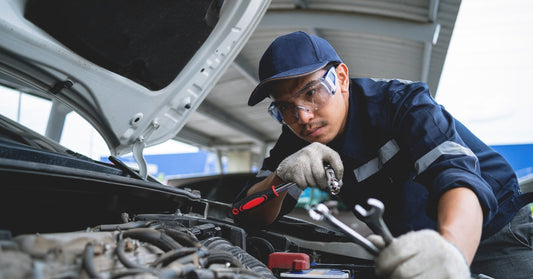 A male mechanic in blue coveralls works on a car engine under an open hood, holding tools, in a bright garage setting.