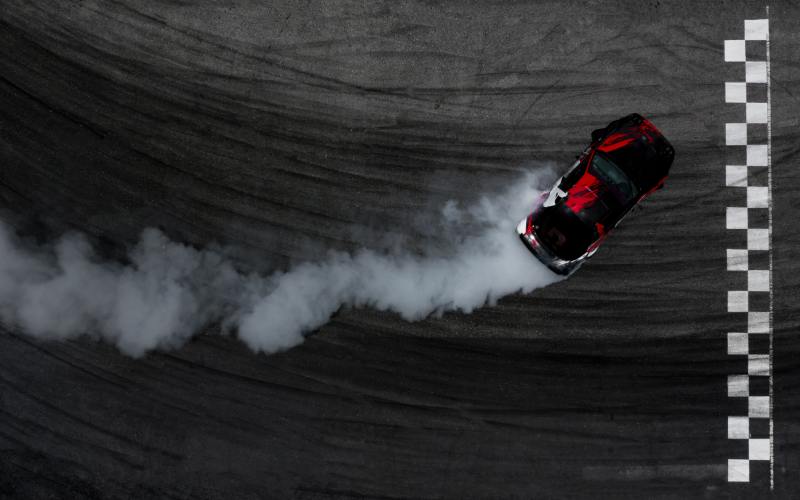 An aerial view of a red and black sports car drifting on a smoky asphalt track near a checkered finish line.