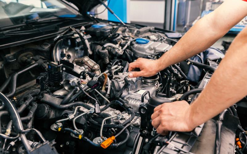 An individual in a red shirt holds a silver wrench above a car engine with hoses, wires, and metal parts.