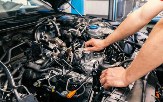 An individual in a red shirt holds a silver wrench above a car engine with hoses, wires, and metal parts.