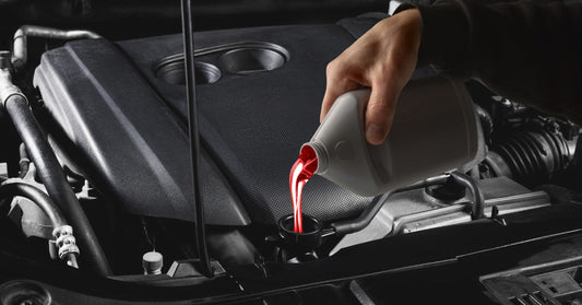 A hand pours bright red liquid from a gray bottle into a car engine with dark hoses and metallic components in view.