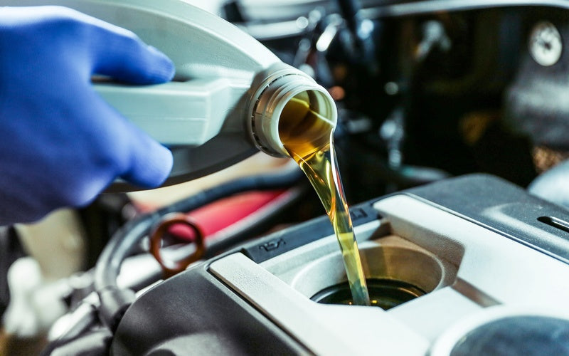 A gloved hand pours golden engine oil from a gray bottle into a car engine, with blurred dark parts in the background.