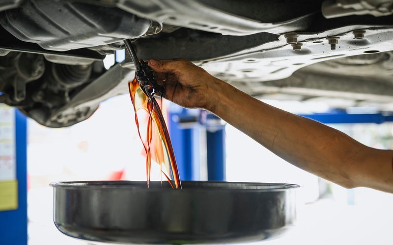 A mechanic's hand uses a wrench to drain amber oil from a car's underside into a black pan in a brightly lit garage.