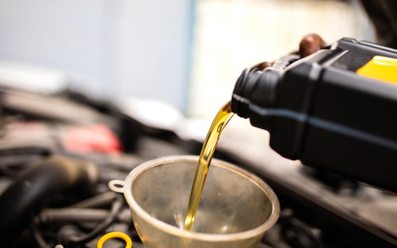 Motor oil poured from a black container into a metal funnel over an open engine bay during a vehicle oil change.