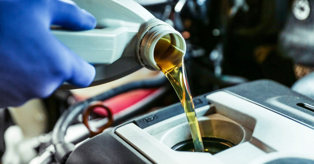 Golden motor oil pours from a white container into a car engine, as a gloved hand holds the bottle over the fill port.