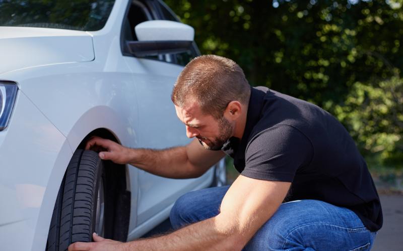 A man crouching beside a white car inspecting the front tire with both hands during an outdoor vehicle check.