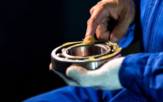 A person applying grease to a metal bearing, with thick lubricant spread across the inner ring against a dark background.