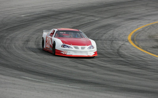 A red and white stock car racing through a banked asphalt turn, hugging the inside line on a worn track surface.