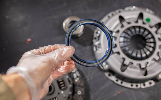 A gloved hand holds a circular blue engine seal ring above clutch components and metal parts on a dark surface.