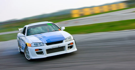 A white race car with a blue stripe drifting through a racetrack corner with green grass and barriers in the background.