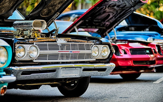 A close-up view of classic hot rods in a variety of bright colors with their hoods open at an outdoor car show.