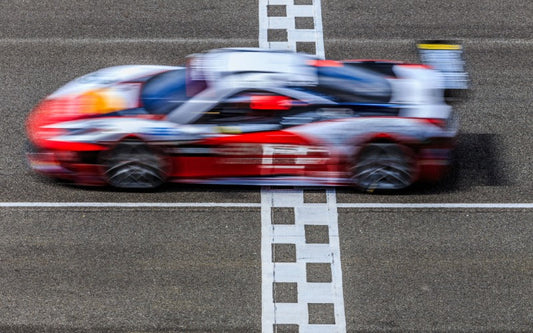 A blurred red and white race car speeds across a painted finish line on an asphalt track during a motorsports race.