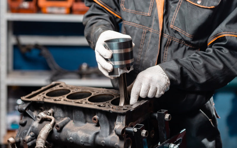 A mechanic installs a piston into an engine block while wearing gloves in a workshop with tools in the background.