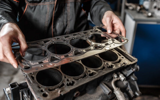 A mechanic positions a metal head gasket over an engine block, aligning cylinder openings during engine assembly.